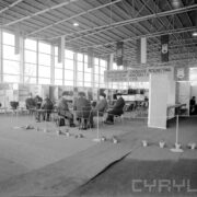 grupa mężczyzn w płaszczach i nakryciach głowy siedzi na krzesłach przy stolikach w hali targowej