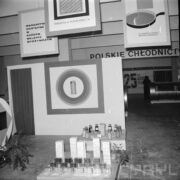 zwisające plakaty reklamujące towary w hali wystawowej, poniżej poukładane butelki i kostki margaryny