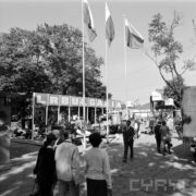 stoisko plenerowe na betonowym placu, zwiedzający ludzie, trzy wysokie maszty z powiewającymi flagami