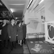 grupa mężczyzna w płaszczach, z prawej kobieta wskazująca ręką na plansze w hali wystawowej