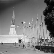 maszty z flagami państw, w głębi okrągły budynek z ażurową iglicą na dachu, na placu spacerujący ludzie