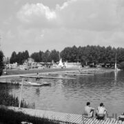 tory regatowe nad jeziorem, w głębi namioty pawilony i spacerujący ludzie, na pierwszym planie drewniany pomost i dwóch siedzących na nim mężczyzn w kąpielówkach