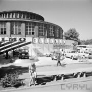 stoisko z samochodami na poboczu ulicy, w głębi murowany okrągły budynek z oknami, przed samochodami spacerujący ludzie, na pierwszym planie ławki bez oparć