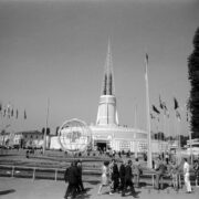 maszty z flagami państw i ażurową konstrukcją kuli ziemskiej pośrodku, w głębi okrągły budynek z ażurową iglicą na dachu, na placu spacerujący ludzie