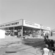 zadaszone stoisko na betonowym placu, na pierwszym planie fragment torów kolejowych, spacerujący ludzie