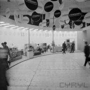 stoisko z maszynami i urządzeniami w hali wystawowej, zwiedzający ludzie, u sufitu wiszą okrągłe reklamy firm