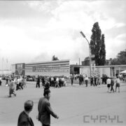 tereny targowe, spacerujący ludzie, w głębi prostokątna reklama z napisem na 25-lecie PRL