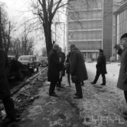 grupa mężczyzn stoi na ośnieżonym chodniku, część podaje sobie dłonie, z lewej stoją zaparkowane samochody