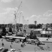 na pierwszym planie aleja na terenie targów, spacerujący ludzie, w głębi budynki wystawowe, i dźwigi budowlane