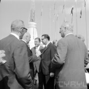 grupa rozmawiających ze sobą mężczyzn w garniturach, za nimi maszty z flagami i budynek z iglicą