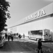 hala wystawowa, przed nią spacerujący ludzie, pod ściana hali zaparkowany bus, w głębi samochody osobowe