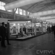 stoiska w hali targowej, pomiędzy nimi chodzą ludzie