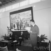 dwóch mężczyzn siedzi przy stoliku, z stoi mężczyzna, w ręce trzyma kartkę papieru, na ścianie duże zdjęcie przywódcy koreańskiego w otoczeniu dzieci