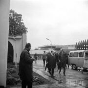 grupa mężczyzn w płaszczach, idących mokrym od deszczu chodnikiem, część trzyma w dłoniach złożone, a część rozłożone parasole