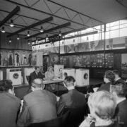 kobieta i mężczyzna siedzą przy stoliku, za nimi stoisko z pralkami, z przodu mężczyźni i kobiety siedzący tyłem