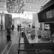 stoiska w hali wystawowej, między nimi zwiedzający ludzie
