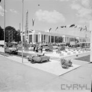 murowany budynek, na pierwszym planie na betonowym placu urządzenia i maszyny, pomiędzy spacerują ludzie, z prawej maszty z flagami państwowymi