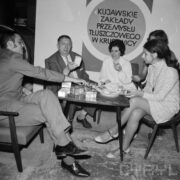 z prawej trzy kobiety, jedna zasłonięta, z lewej dwaj mężczyźni, wszyscy siedzą na fotelach przy małym stoliku