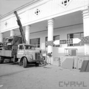 samochód z dźwigiem, na jego przyczepie maszyna, za samochodem fragment otynkowanego budynku z kolumnami