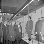 grupa mężczyzn, część w płaszczach, ogląda ubrane manekiny w hali wystawowej