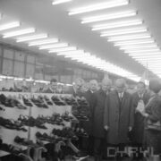 grupa ludzi, większość w płaszczach w hali wystawowej, po lewej półka z butami