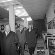 grupa ludzi, część w płaszczach, w hali wystawowej