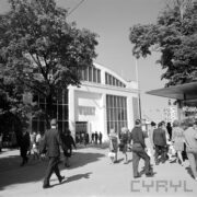 murowany budynek, z przeszkleniami, przed nim ludzie, po prawej i po lewej stronie drzewa