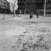 pusty plac przecięty deptakiem, na nim przechodnie, w głębi kamienice