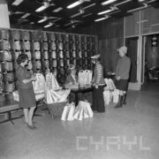 przy stoliku stoją trzy osoby, pośrodku siedzi jedna kobieta, wokół stolika i przy nim ustawiono stoi kilkadziesiąt papierowych toreb