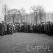 duża grupa ludzi w płaszczach i nakryciach głowy (w większości) stoi w półkolu, za nimi pawilon wystawowy