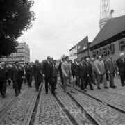 duża grupa ludzi idzie po brukowanej alei na terenie targów, w głębi budynki targowe