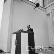 mężczyzna stoi przy mównicy przyozdobionej polską flagą z orłem i przemawia