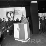 pośrodku mównica przybrana godłem państwowym oraz barwami polskiej flagi, na mównicy przemawia mężczyzna, w głębi, po obu jej stronach stoją mężczyźni