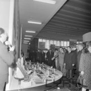 grupa mężczyzn, część w płaszczach, część w garniturach i kilka kobiet oglądają towary wystawione na stoiskach w hali targowej