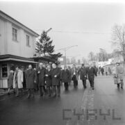 grupa mężczyzn w płaszczach idzie wewnętrzną drogą na tragach, z lewej fragment pawilonu targowego