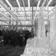 grupa mężczyzn w płaszczach, część w garniturach, w hali wystawowej ogląda stoisko z tkaninami