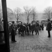 duża grupa mężczyzn w płaszczach, kilku trzyma w rękach bukiety kwiatów opakowane w celofan, stoi na chodniku przed pawilonem wystawowym