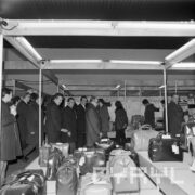 grupa mężczyzn w płaszczach ogląda ekspozycję skórzanych toreb w hali targowej