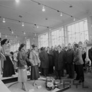 grupa mężczyzn w płaszczach, część w garniturach, w hali wystawowej ogląda odzież prezentowaną na manekinach