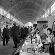 grupa mężczyzn w płaszczach w hali wystawowej ogląda stoisko z odzieżą prezentowaną na manekinach