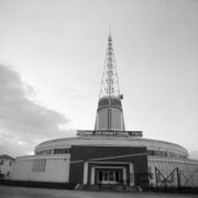 okrągły budynek z ażurową iglicą na dachu, nad wejściem do pawilonu świetlny napis