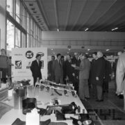 grupa mężczyzn, część w płaszczach, część w garniturach ogląda ekspozycje w hali targowej