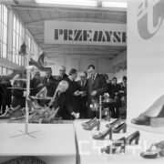 grupa mężczyzn w płaszczach, jeden w garniturze, w hali wystawowej ogląda stoisko z butami