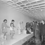 grupa mężczyzn w płaszczach w hali wystawowej ogląda stoisko z odzieżą prezentowaną na dziecięcych manekinach