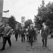 grupa mężczyzn idzie aleją na terenie targów, w głębi, z lewej pawilon wystawienniczy, z lewej mężczyzna w mundurze milicjanta