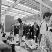 grupa mężczyzn w płaszczach w hali wystawowej ogląda stoisko z odzieżą prezentowaną na dziecięcych manekinach