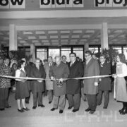 grupa mężczyzn w płaszczach stoi przed szarfą, podtrzymywaną z prawej strony przez młodą kobietę, pośrodku mężczyzna przecina szarfę nożyczkami