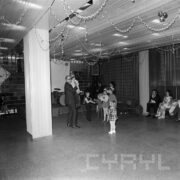 mężczyzna stoi z mikrofonem, na ręce trzyma dziecko, przy nim stoi kilkoro dzieci, z sufitu zwisają serpentyny