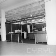 stoisko w hali wystawowej, przy nim, w głębi dwóch mężczyzn