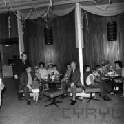 grupa kobiet i mężczyzn z dziećmi siedzi przy małych stolikach, na których przygotowano poczęstunek